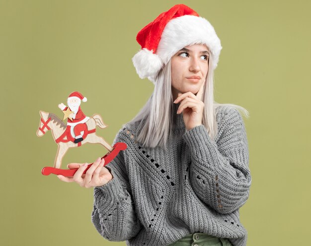 Young blonde woman in winter sweater and santa hat holding christmas toy looking aside puzzled standing over green wall
