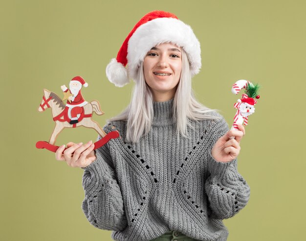 Молодая блондинка в зимнем свитере и шляпе санта-клауса с елочными игрушками, весело улыбаясь, стоя у зеленой стены