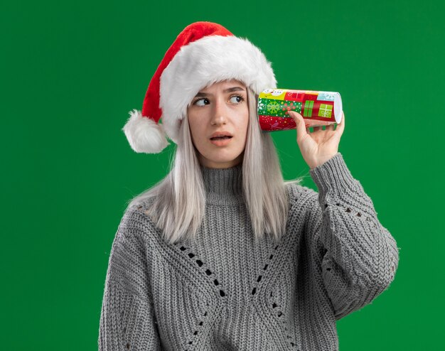 Young blonde woman in winter sweater and santa hat holding colorful paper cup  over her ear looking intrigued standing over green background