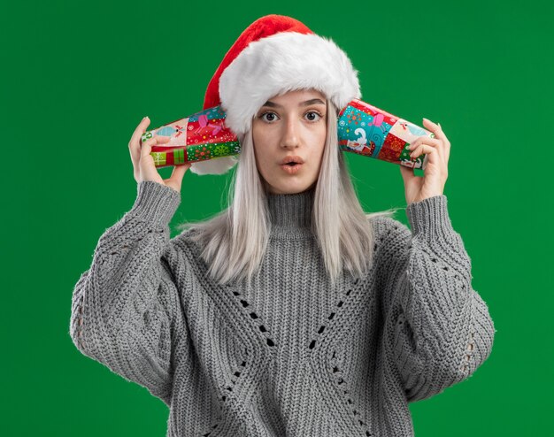 Young blonde woman in winter sweater and santa hat holding colorful paper cups over her ears looking confused  standing over green background