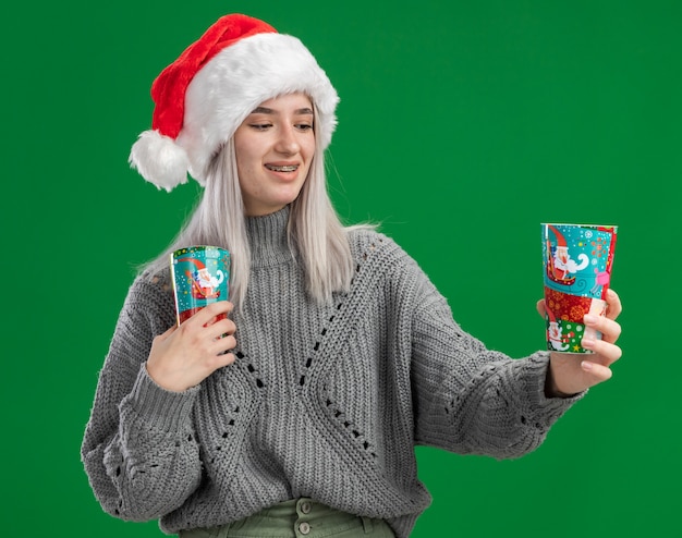 Young blonde woman in winter sweater and santa hat holding colorful paper cups looking at cup smiling with happy face  standing over green background