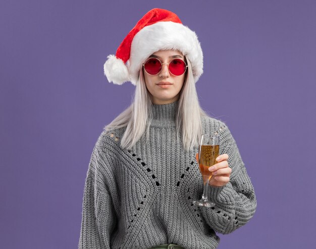 Молодая блондинка в зимнем свитере и шляпе санта-клауса держит бокал шампанского с уверенным выражением лица, стоя над фиолетовой стеной