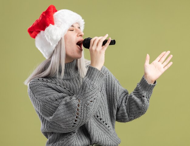 Giovane donna bionda in maglione invernale e cappello santa tenendo il microfono cantando felice e positivo che celebra la festa di natale