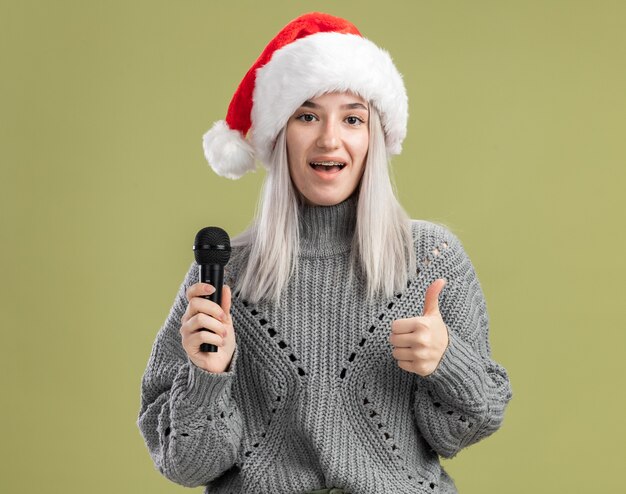 Giovane donna bionda in maglione invernale e cappello da babbo natale che tiene il microfono con un sorriso sul viso che mostra i pollici in piedi sul muro verde