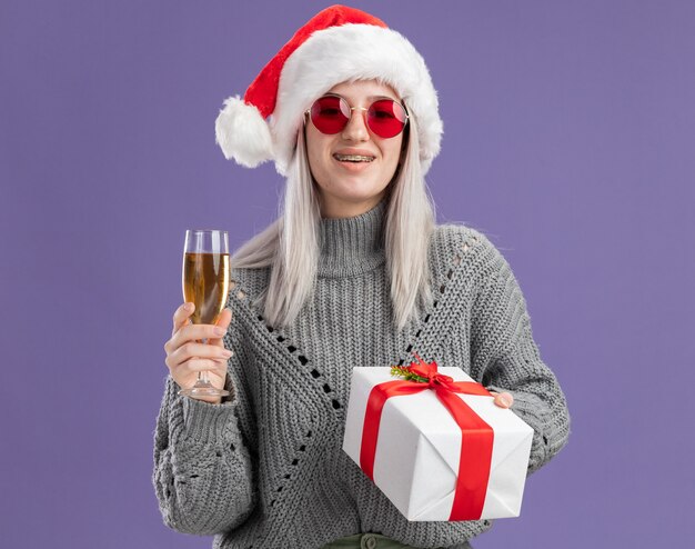 Молодая блондинка в зимнем свитере и шляпе санта-клауса держит подарок и бокал шампанского с улыбкой на счастливом лице, стоящем над фиолетовой стеной
