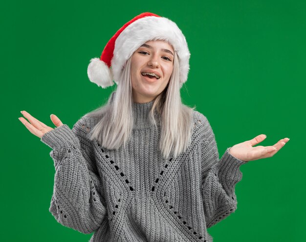 겨울 스웨터와 산타 모자에 젊은 금발의 여자 행복하고 긍정적 인 녹색 배경 위에 유쾌하게 서 웃고