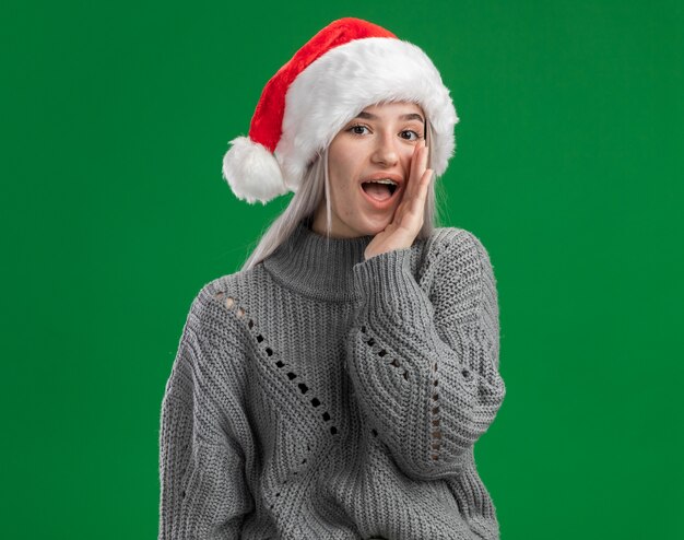 Young blonde woman in winter sweater and santa hat telling a secret whispering with hand near mouth  standing over green background