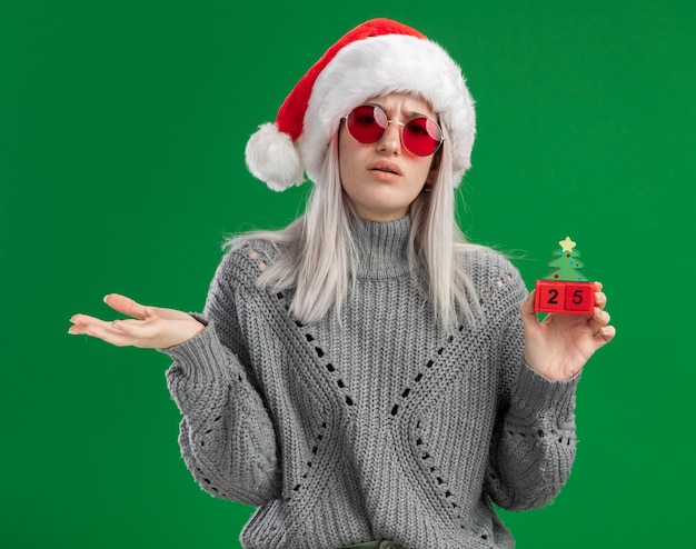 Giovane donna bionda in maglione invernale e santa hat indossando occhiali rossi tenendo i cubi giocattolo con la data di natale cercando confuso con il braccio in piedi su sfondo verde