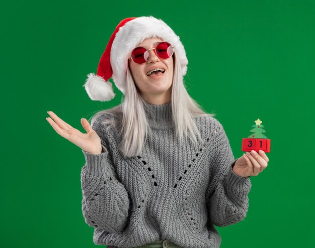 겨울 스웨터와 산타 모자에 젊은 금발의 여자 해피 뉴 이어 날짜 행복하고 긍정적 인 녹색 배경 위에 유쾌하게 서있는 장난감 큐브를 들고 빨간 안경을 쓰고