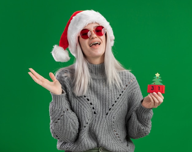 Young blonde woman in winter sweater and santa hat wearing red glasses holding toy cubes with happy new year date happy and positive smiling cheerfully  standing over green background