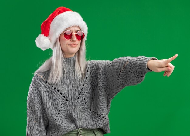 Young blonde woman in winter sweater and santa hat wearing red glasses  looking aside with serious face pointing with index finger at something standing over green background