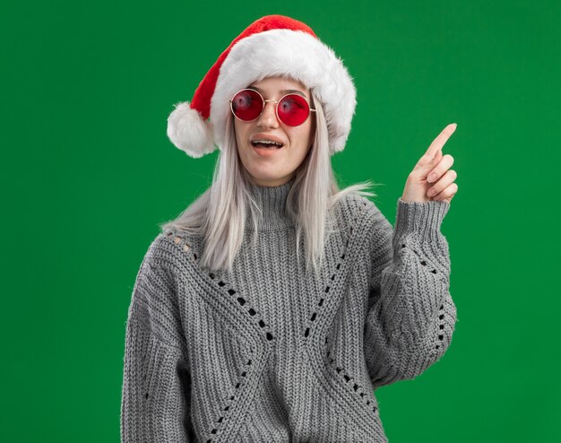 Giovane donna bionda in maglione invernale e cappello da babbo natale con gli occhiali rossi che sembrano sorpresi mostrando il dito indice con una nuova idea in piedi su sfondo verde