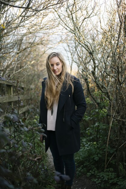 Young blonde woman with a black coat standing on a pathway surrounded by leafless trees