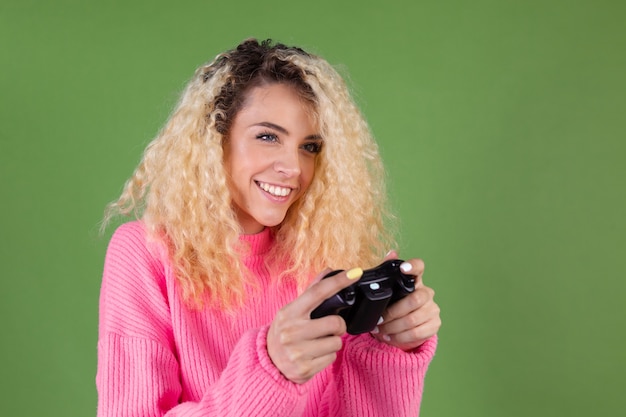 Young blonde woman with long curly hair in pink sweater on green with joystick playing games