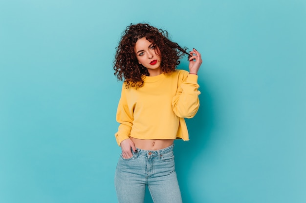 Young blue-eyed woman with red lips coquettishly touches curly hair and poses in yellow-blue outfit on isolated space.