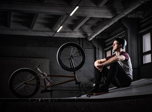 Young Bmx rider relaxing after practicing tricks with his bike in a skatepark indoors