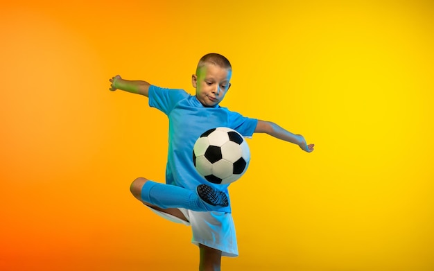 Young boy as a soccer player in sportwear practicing on gradient yellow studio background in neon light
