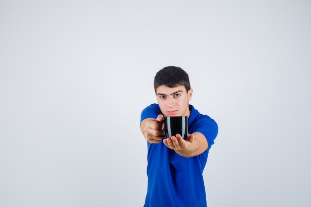 Free photo young boy in blue t-shirt holding cup, giving it to someone and looking confident , front view.