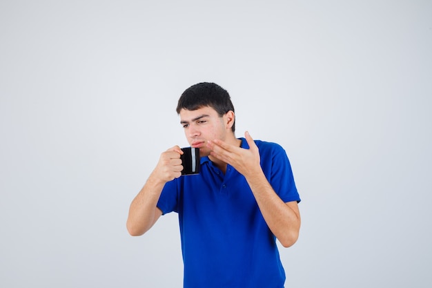 Молодой мальчик в синей футболке держит чашку, пытается пить из нее жидкость и смотрит сосредоточенно, вид спереди.