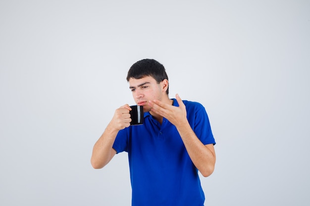 Young boy in blue t-shirt holding cup, trying to drink liquid from it and looking focused , front view.