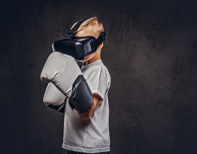 Free photo young boy boxer with blonde hair dressed in a white t-shirt wearing visual reality glasses and boxing gloves, workout in a studio. isolated on a dark textured background.