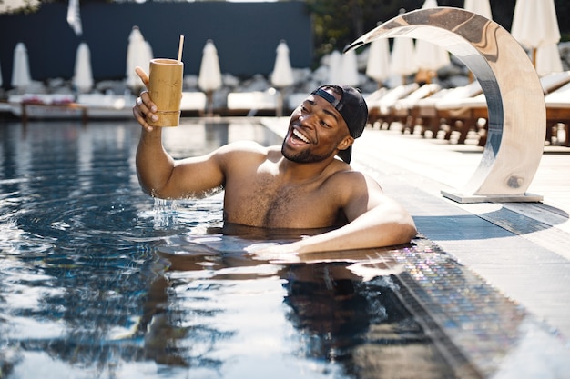 Ragazzo in un berretto in piedi in una piscina e ha un cocktail. uomo che beve un cocktail da un bicchiere di bambù.