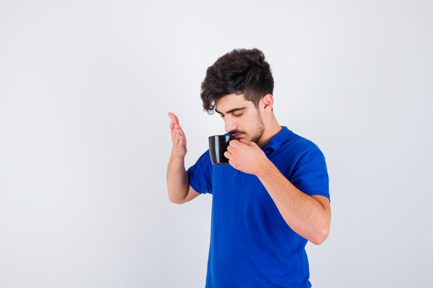 Young boy drinking cup of tea and stretching hand in blue t-shirt and looking serious , front view.