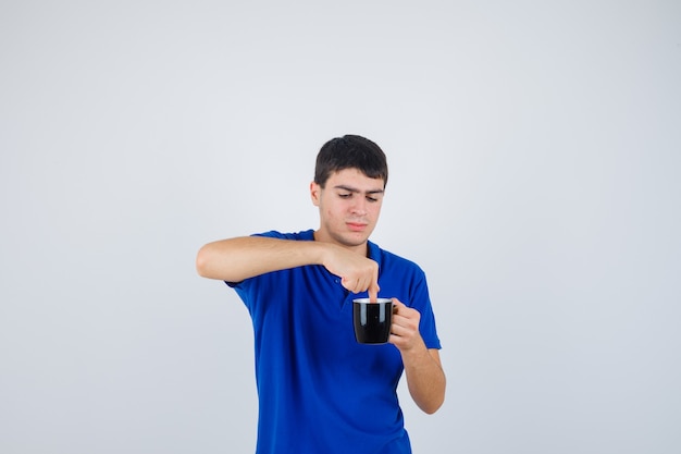Мальчик держит чашку возле подбородка, кладет в нее руку в синей футболке и смотрит с любопытством, вид спереди.