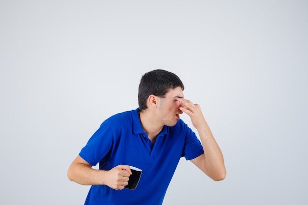 Young boy holding cup, pinching nose due to bad smell in blue t-shirt and looking irritated. front view.