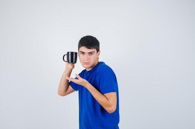 Young boy holding cup, stretching hand as presenting it in blue t-shirt and looking serious. front view.