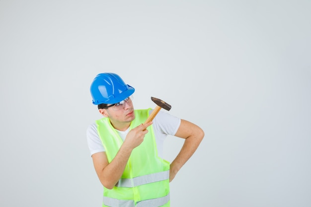 Young boy holding hammer, putting hand on waist, looking at hammer in construction uniform and looking focused. front view.