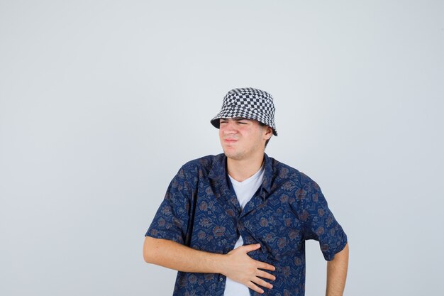 Young boy holding hand on belly, having bellyache in white t-shirt, floral shirt, cap and looking exhausted