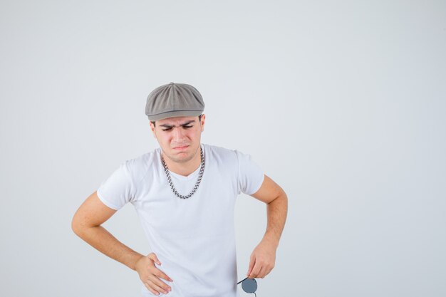 Young boy holding hand on waist in t-shirt, hat and looking painful. front view.