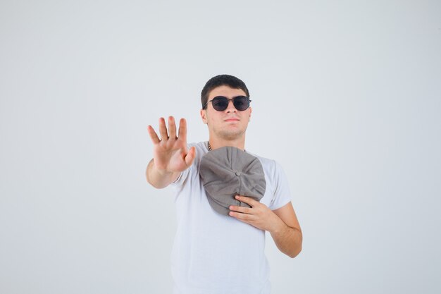 Young boy holding hat on chest while showing palm in t-shirt and looking confident , front view.