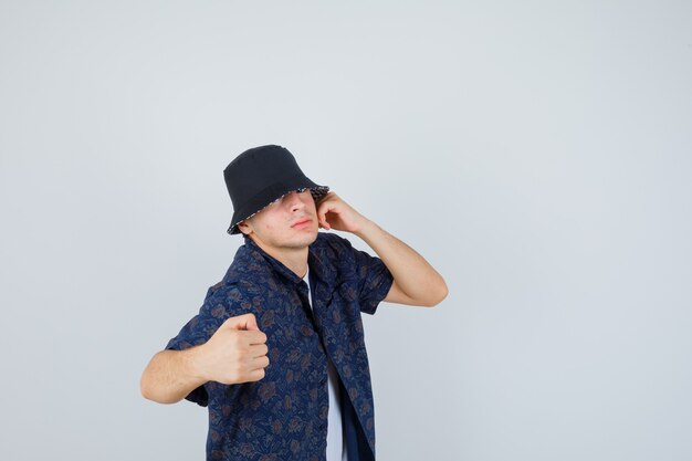 Young boy holding one hand near ear, clenching fist while posing in white t-shirt, floral shirt, cap and looking confident , front view.