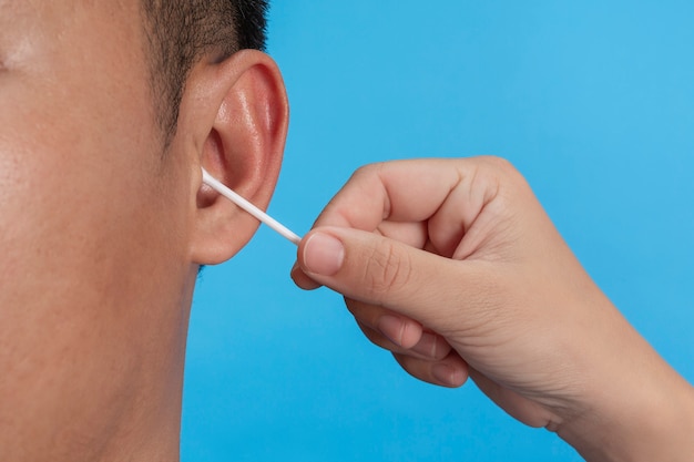 Young boy is ear picking with cotton swab on blue wall, studio.