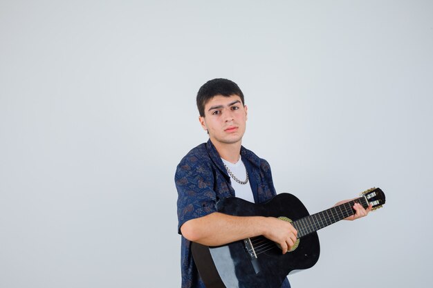 Young boy playing guiter while sitting aganist, looking at camera in t-shirt and looking confident. front view.