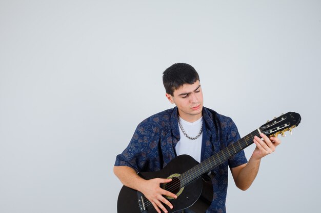 Young boy playing guiter while sitting aganist in t-shirt and looking confident , front view.