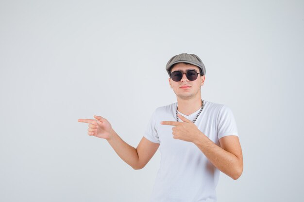 Young boy pointing to the left side in t-shirt, hat and looking self-confident. front view.