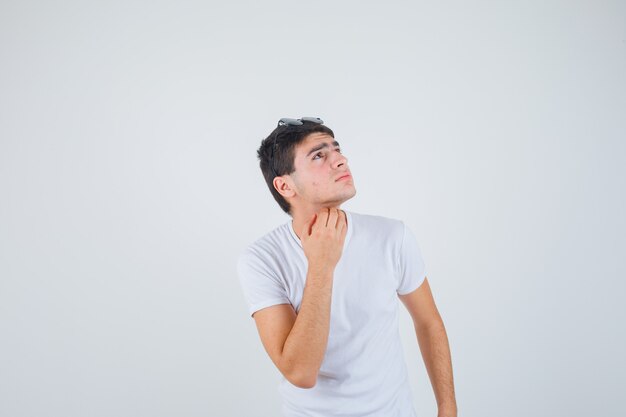 Young boy scratching throat while looking upward in t-shirt and looking pensive , front view.