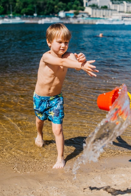 Young boy in shorts throwing bucket with water on sea beach
