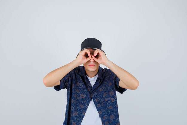 Young boy showing binoculars gesture in white t-shirt, floral shirt, cap and looking serious.