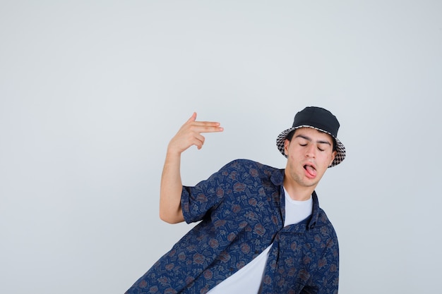 Free photo young boy showing gun gesture near head, sticking tongue out in white t-shirt, floral shirt, cap and looking confident