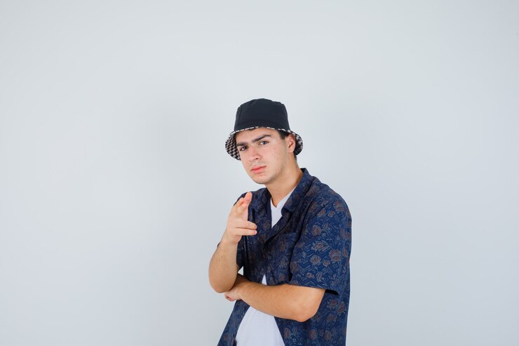 Free Photo young boy showing gun gesture toward camera, holding hand under elbow in white t-shirt, floral shirt, cap and looking confident