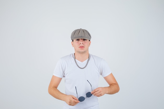 Молодой мальчик в футболке, шляпе держит очки, смотрит в камеру и выглядит уверенно, вид спереди.