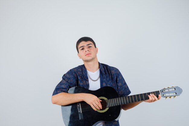 Young boy in t-shirt playing guiter while sitting aganist and looking confident , front view.