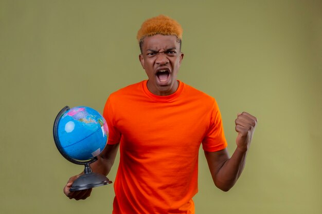Young boy wearing orange t-shirt holding globe clenching fist shouting with angry expression on face standing over green wall