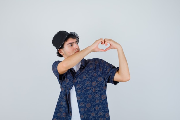 Young boy in white t-shirt, floral shirt, cap showing heart gesture and looking confident