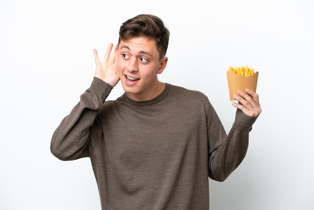 Young brazilian man holding fried chips isolated on white background listening to something by putting hand on the ear