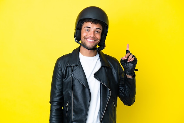 最高の兆候を示して指を持ち上げて黄色の背景に分離されたオートバイのヘルメットを持つ若いブラジル人男性